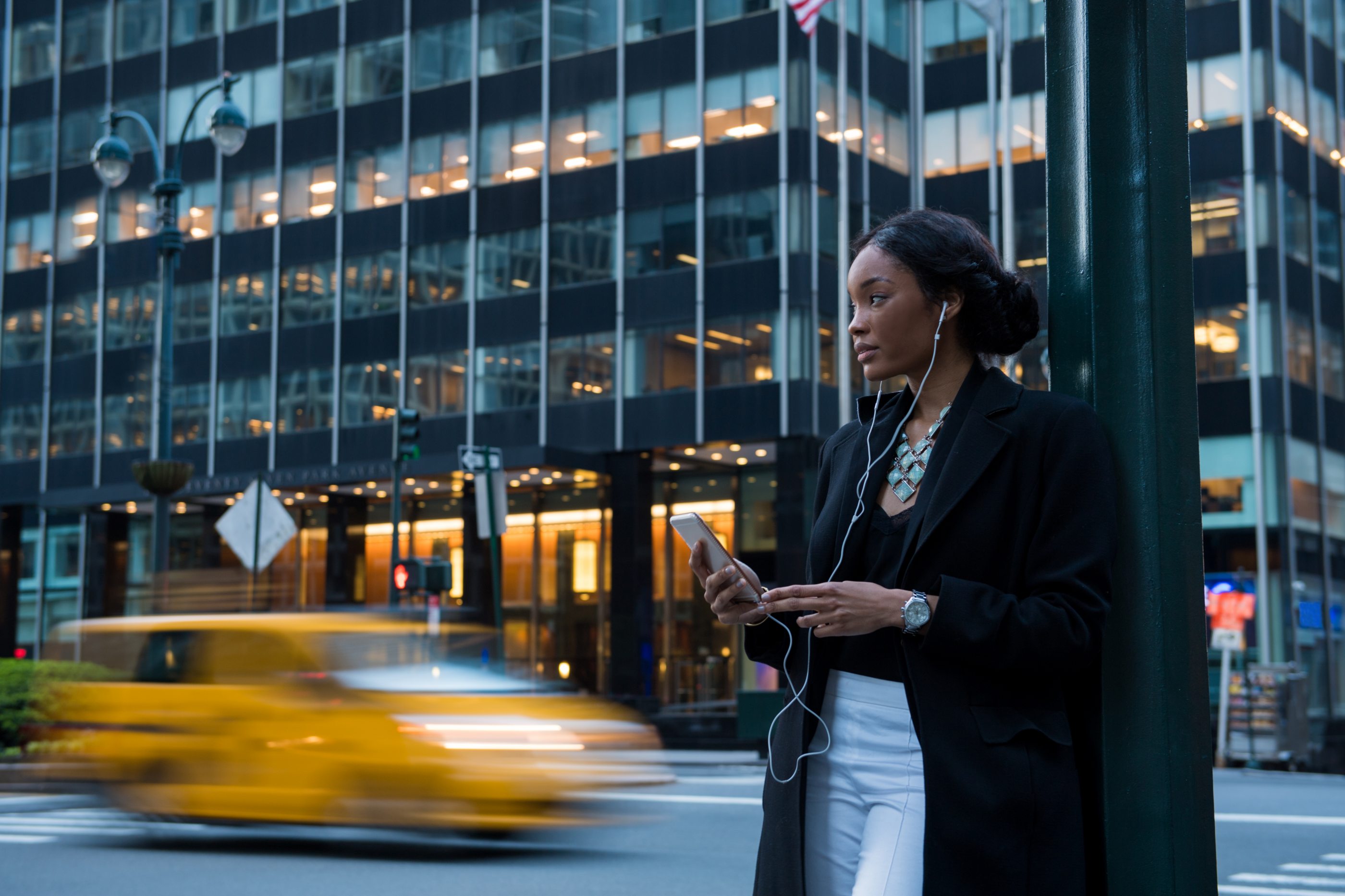 A woman with earphone from a cellphone standing against a pole next to a street with cars passing