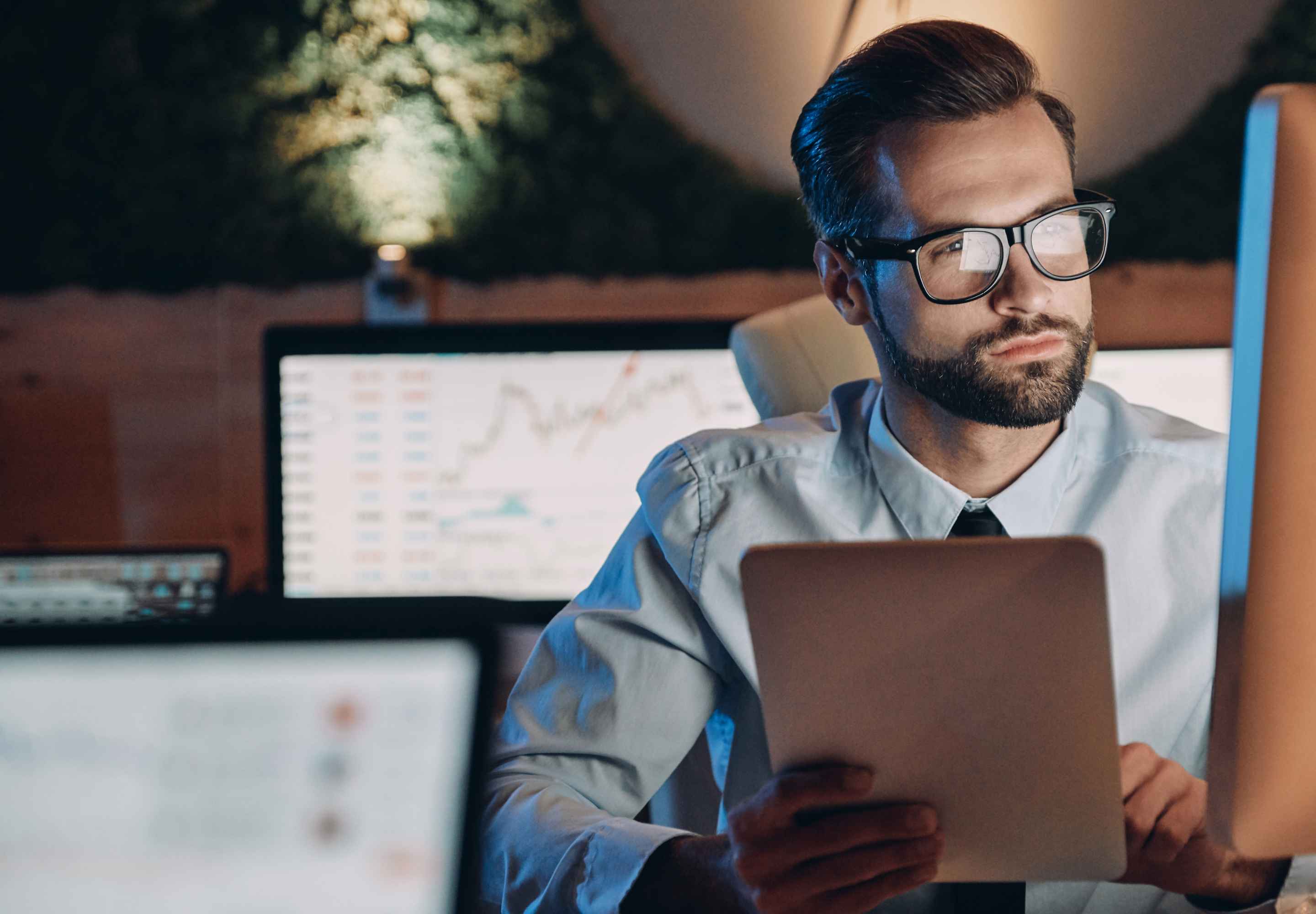 A man holding tablet device while looking at big monitor next to laptop