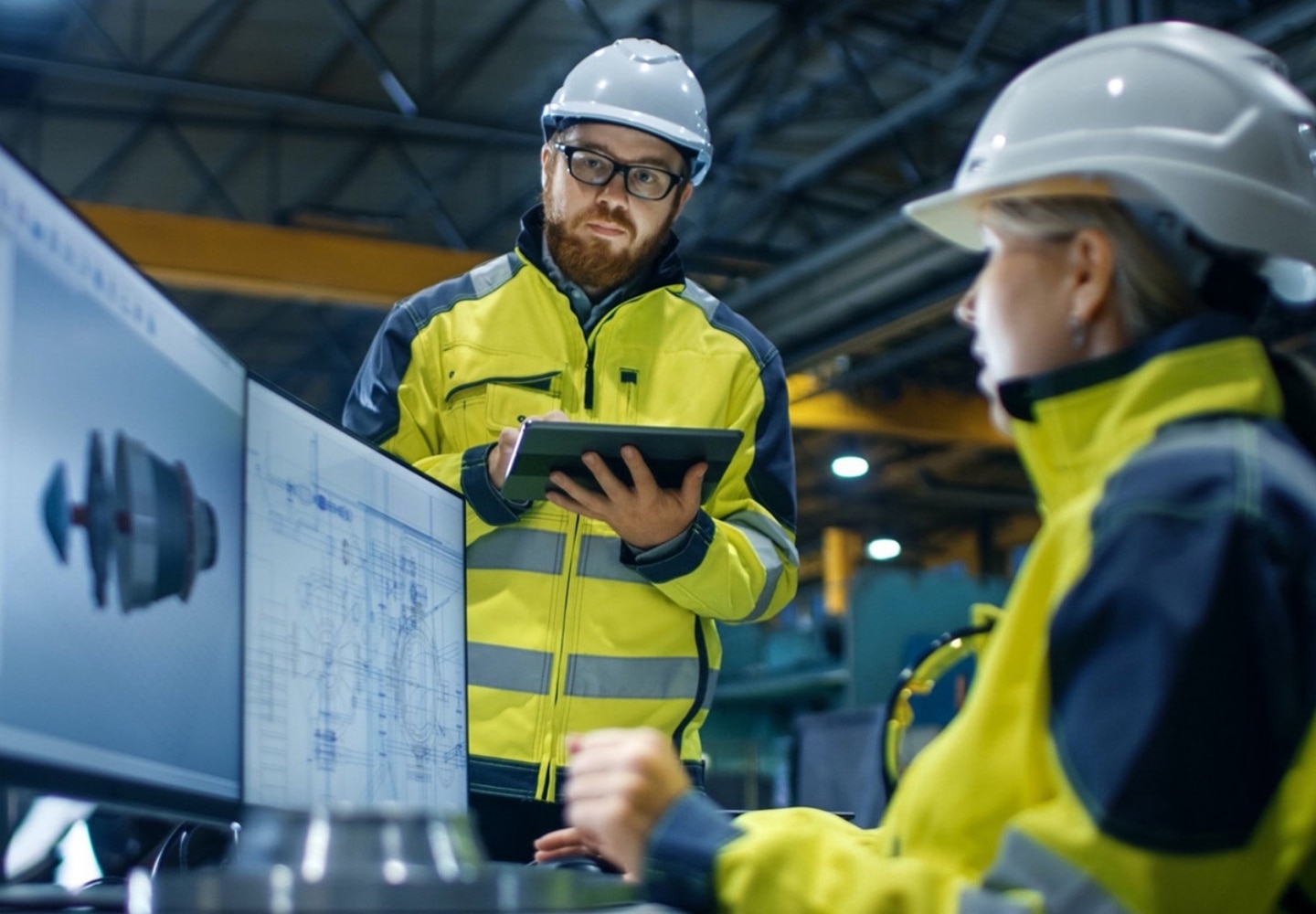 A man and a woman in construction wear looking at two monitor screens