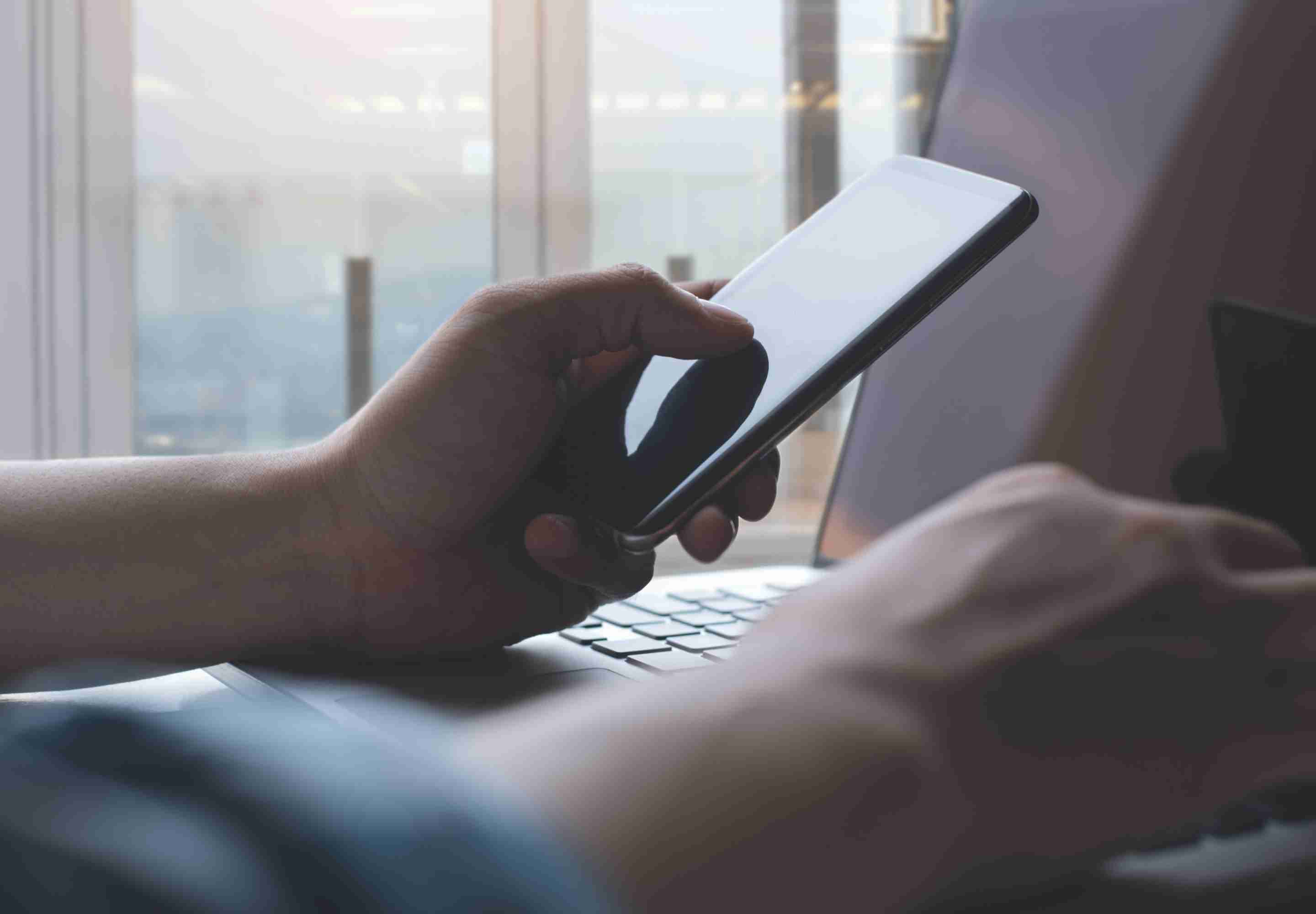 A person holding a cell phone while typing on a laptop