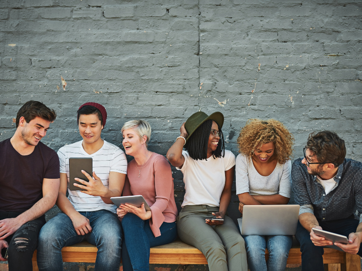 A group of people sitting on a bench with various laptops and tablets
